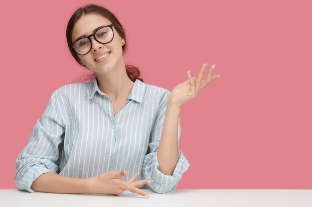 Foto gratuita mujer europea joven atractiva de moda en camisa azul formal y anteojos sentado en la mesa aislada, sonriendo ampliamente, haciendo gesto amistoso de bienvenida, posando en la pared de copyspace rosa en blanco