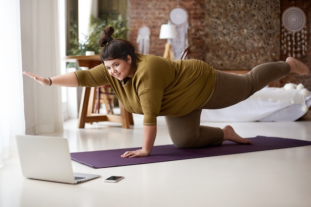 Mujer europea joven gordita obesa con nudo de pelo practicando yoga o pilates en el interior sobre una colchoneta, haciendo ejercicios para fortalecer el núcleo, viendo una lección de video en línea frente a una computadora portátil abierta en el piso