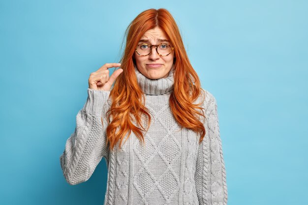 Mujer europea joven pelirroja disgustada vestida con un jersey de invierno gris muestra gesto de pequeña cantidad demuestra tamaño pequeño con los dedos.