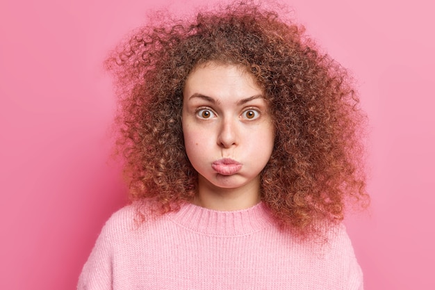 La mujer europea joven de pelo rizado ofendida sopla las mejillas hace muecas vestida con un jersey casual contiene la respiración parece sorprendentemente aislada sobre la pared rosada. Concepto de expresiones faciales