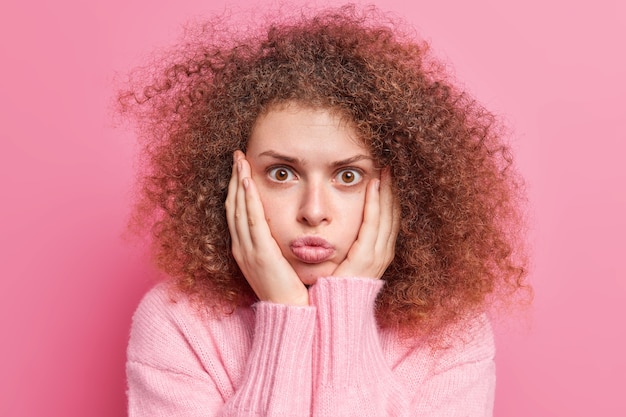 La mujer europea joven de pelo rizado sorprendida mantiene las manos en las mejillas mira fijamente los labios conmocionados pucheros nota algo increíble vestido casualmente aislado sobre la pared rosada. Concepto de Omg