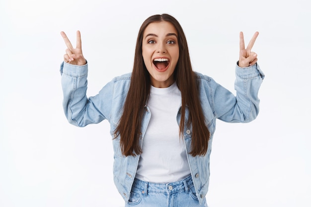 Foto gratuita mujer europea linda positiva y optimista en chaqueta de mezclilla, jeans, mostrando signos de paz o victoria, haciendo comillas en el aire y sonriendo como sarcástico o regañando a alguien, de pie fondo blanco