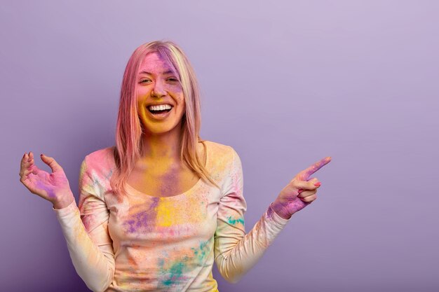 La mujer europea llena de alegría se ríe de las impresiones positivas, muestra el lugar donde se lleva a cabo el festival de Holi, se divierte con polvos de colores, untado con tintes de colores, sonríe ampliamente. Celebración en India