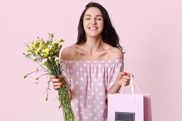 Una mujer europea morena contenta sostiene una bolsa de regalo y flores, feliz de recibir el regalo en el Día de la Mujer, vestida con un vestido de lunares, muestra los hombros desnudos, tiene un aspecto positivo, posa en interiores.