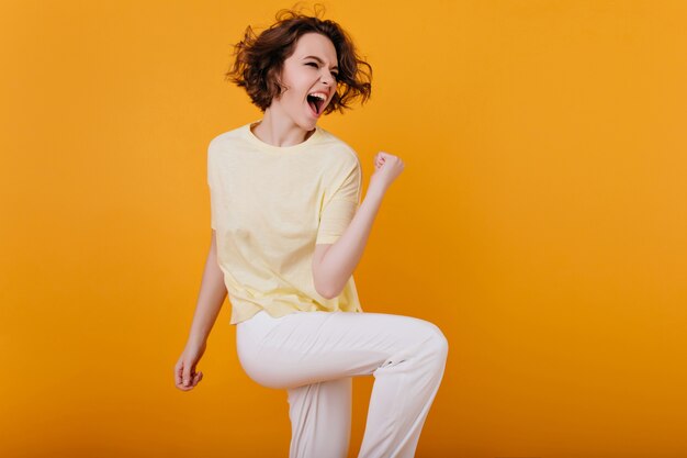 Mujer europea muy emocionada divertida bailando con interior naranja. Foto interior de niña rizada entusiasta en traje blanco pasar tiempo en casa.