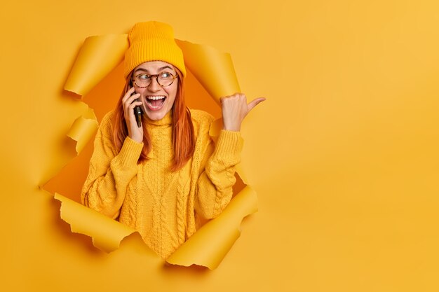 La mujer europea pelirroja alegre feliz lleva sombrero y un suéter caliente tiene una charla positiva a través de los puntos del teléfono inteligente en el espacio en blanco.