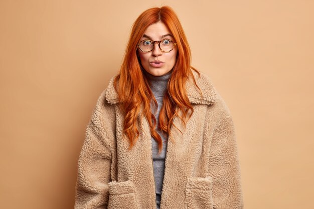La mujer europea pelirroja asombrada oye rumores increíbles o impresionado por algo secreto lleva gafas transparentes y abrigo marrón de piel.