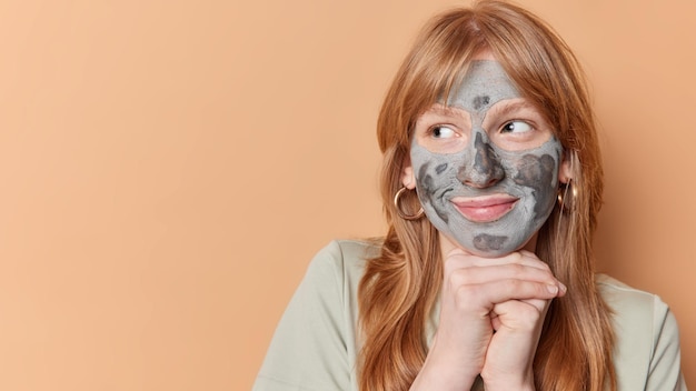 Foto gratuita una mujer europea pelirroja complacida mantiene las manos bajo la barbilla y mira hacia otro lado aplica una máscara de belleza de arcilla en la cara vestida casualmente aislada sobre un espacio de copia de fondo marrón para colocar su anuncio