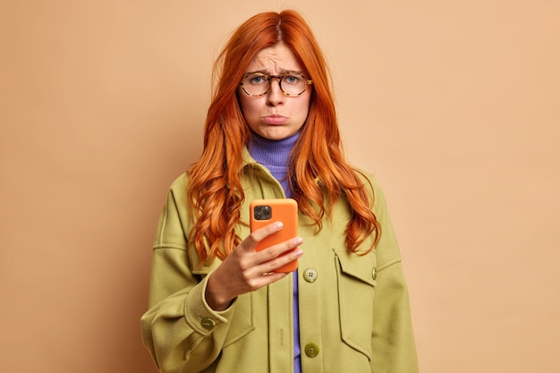 Foto gratuita mujer europea pelirroja disgustada frustrada molesta porque su novio no llama a los puestos ofendido utiliza el teléfono móvil para navegar por internet y lleva una chaqueta verde.