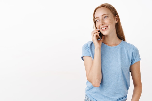 Mujer europea pelirroja ordinaria relajada despreocupada con pecas en camiseta azul girando a la izquierda mientras sostiene el teléfono cerca de la oreja y habla