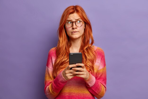 La mujer europea pelirroja preocupada piensa qué respuesta dar en el mensaje recibido recientemente lleva gafas ópticas y un suéter concentrado a un lado con una expresión dudosa.