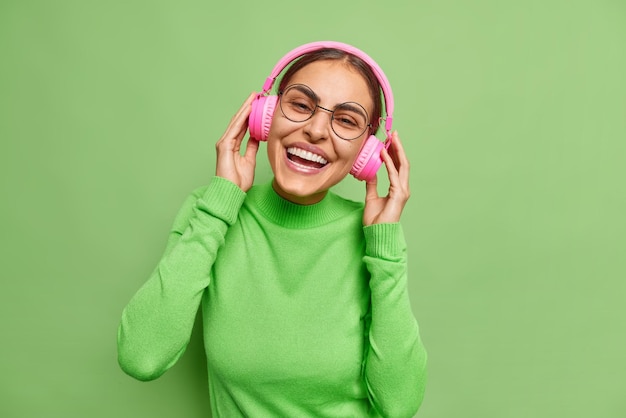 Una mujer europea de pelo oscuro positiva mantiene las manos en los auriculares estéreo rosas y disfruta escuchando música sonríe con dientes vestida con un cuello de tortuga casual aislado sobre fondo verde Toma monocromática
