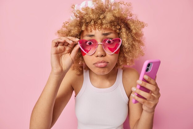 Mujer europea de pelo rizado insatisfecha mira la cámara a través de gafas de sol rosadas no puede creer que sus propios ojos apliquen parches de belleza debajo de los ojos sostiene el teléfono móvil vestido con una camiseta posa en el interior