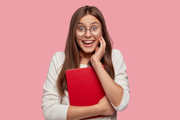 Mujer europea positiva con cabello lacio, tiene una sonrisa dentuda, se ríe mientras se divierte con sus compañeros de clase, usa gafas redondas, sostiene un libro de texto rojo, se regocija al recibir una buena nota, modelos sobre una pared rosa