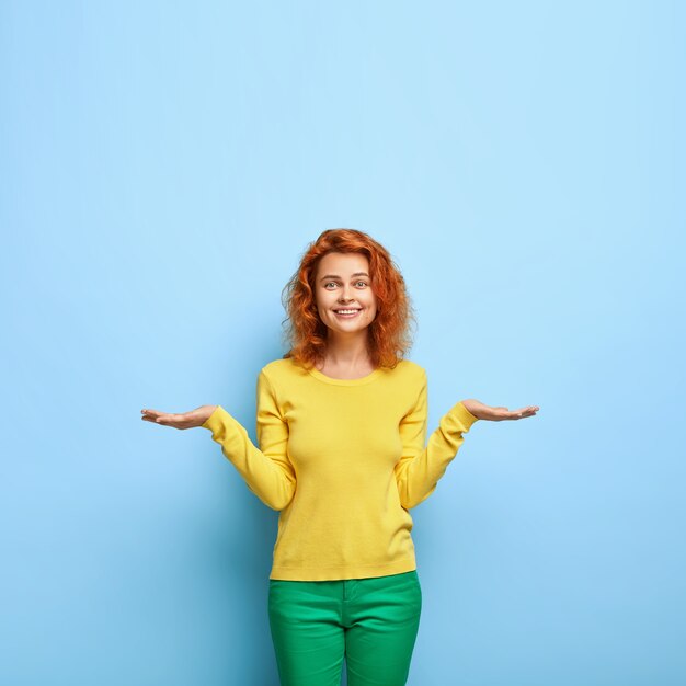 Mujer europea positiva complacida con peinado de jengibre levanta dos palmas como si sosteniendo algo hiciera elegir entre dos objetos invisibles vestidos con sudadera amarilla que se encuentra sobre un espacio en blanco de la pared azul