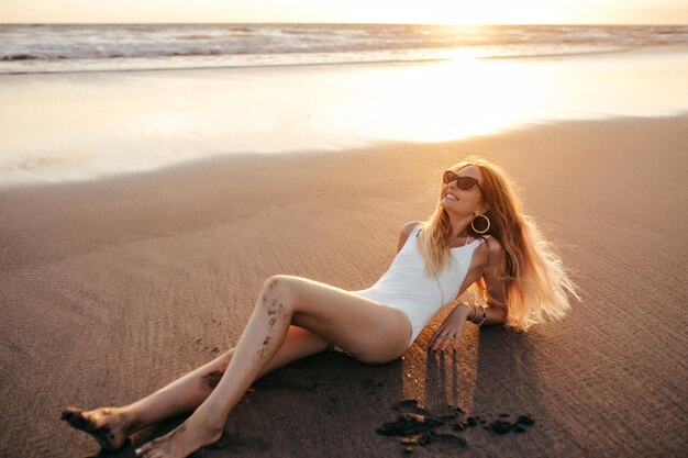 Mujer europea positiva en elegante traje de baño tumbado en la arena y mirando al sol Retrato al aire libre de una chica alegre con el pelo largo tomando el sol en un complejo exótico