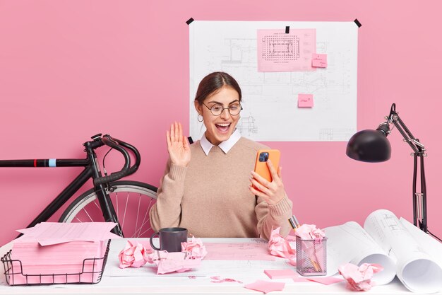 La mujer europea positiva hace la palma de la mano de las ondas de la videollamada, sostiene el teléfono inteligente moderno trabaja en el informe prepara las poses de la tarea en el hogar