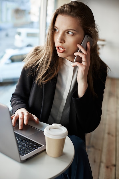 Mujer europea preocupada seria sentada en la cafetería, tomando café y trabajando con la computadora portátil, hablando por teléfono inteligente mientras mira a un lado con ansiedad