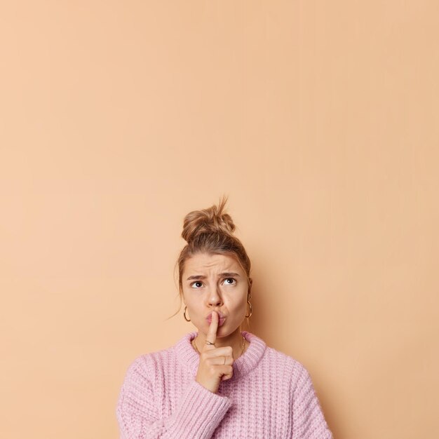 Una mujer europea seria y enfadada hace un gesto de silencio que mantiene el dedo índice sobre los labios concentrado sobre la cara fruncida usa un puente casual aislado sobre un espacio de copia de fondo beige para su promoción