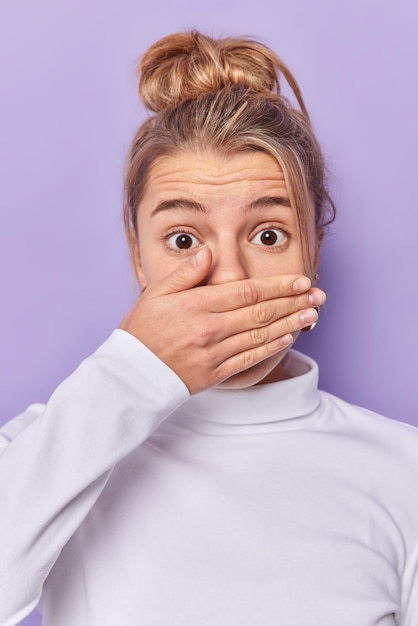 Foto gratuita una mujer europea sorprendida y aturdida se cubre la boca con la palma de la mano, se ve sorprendida, trata de quedarse sin palabras, usa un jersey blanco casual, se siente estupefacta, aislada sobre un fondo morado. concepto de reacciones humanas.