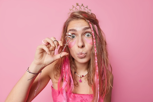 La mujer europea sorprendida se prepara para la fiesta usa el rizador de pestañas aplica parches de hidrogel de belleza de maquillaje debajo de los ojos mantiene los labios redondeados usa corona y vestido festivo aislado sobre fondo rosa.