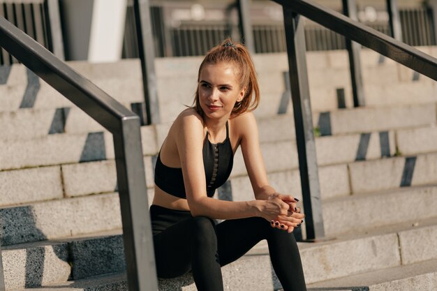 Mujer europea en uniforme deportivo sentado en las escaleras y preparándose para el entrenamiento exterior