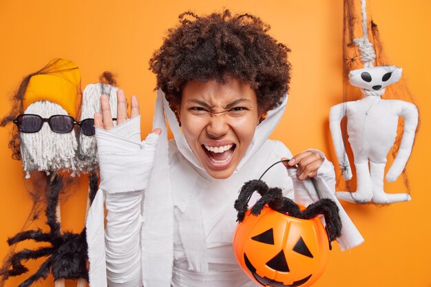 Mujer exclama en voz alta sostiene calabaza tallada con araña vestida como momia para posturas de fiesta de Halloween en naranja sobre decoraciones tradicionales cuenta historias de miedo