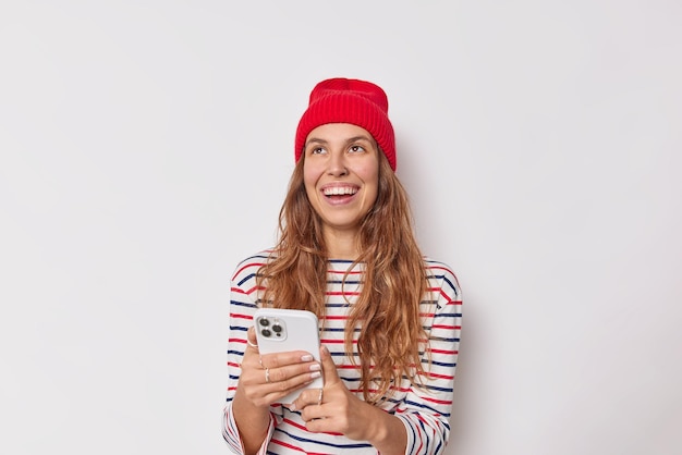 mujer con expresión feliz usa gadget de teléfono móvil viste un puente de rayas casual de sombrero rojo aislado en blanco. Personas tecnologías modernas y concepto de estilo de vida.