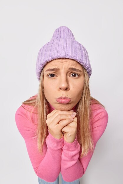 Mujer con expresión de lástima bolsas de labios tiene expresión suplicante viste gorro de punto y jersey rosa aislado en blanco