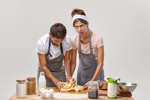 La mujer fatiga le enseña al esposo a hacer masa, explica cómo amasar y qué ingredientes agregar, preparar la cena festiva, hornear en casa, rodeada de los productos necesarios en la mesa, probar una nueva receta