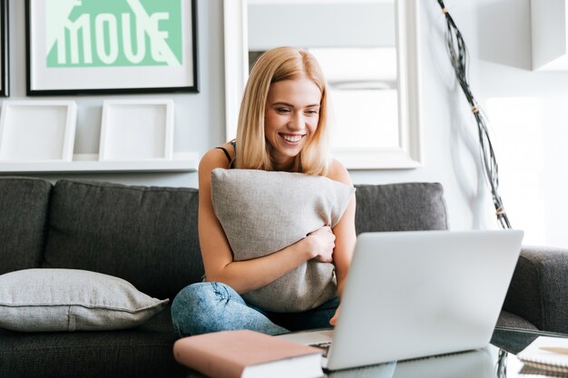 Mujer feliz abrazando la almohada y usando la computadora portátil en el sofá