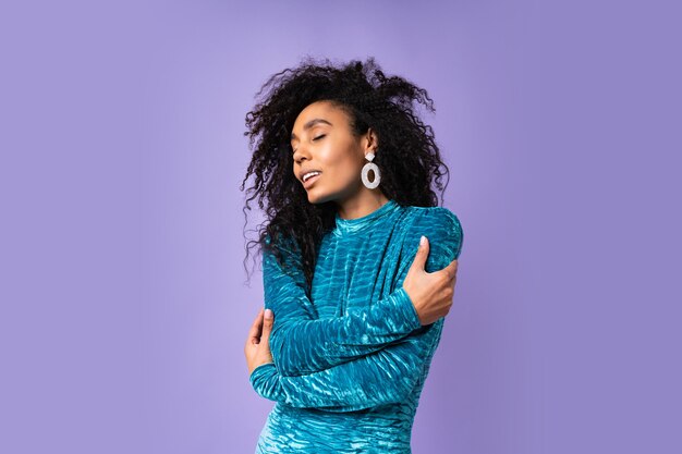 Mujer feliz afroamericana con los ojos cerrados en vestido de terciopelo con cabello ondulado posando. Retrato de estilo de moda.