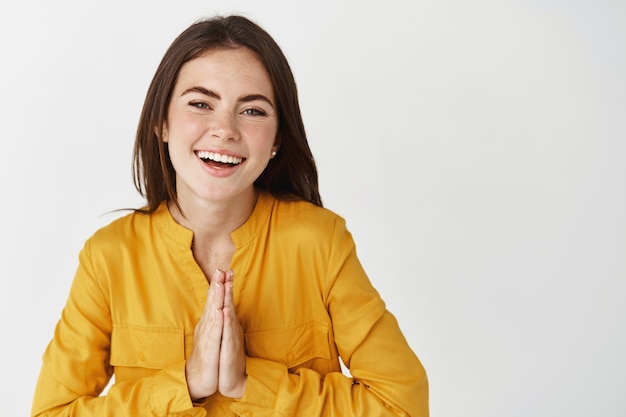 Mujer feliz y agradecida diciendo gracias, sonriendo con gratitud, juntando las manos en el pecho, de pie sobre la pared blanca