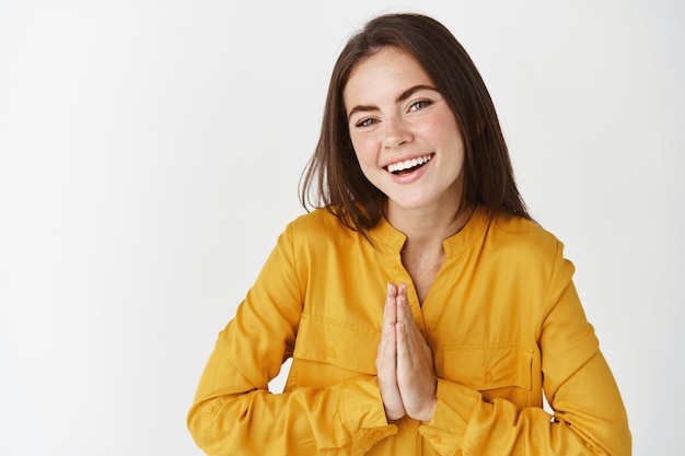 Mujer feliz y agradecida diciendo gracias, sonriendo con gratitud, juntándose las manos en gesto de namaste, de pie sobre una pared blanca