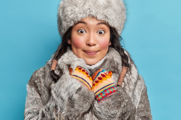 Foto gratuita mujer feliz de alaska mira con cara sorprendida y encantada en la parte delantera lleva sombrero de invierno abrigo de piel y mitones posa contra la pared azul