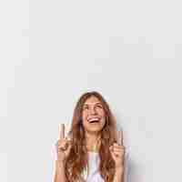 Foto gratuita la mujer feliz y alegre con el pelo largo indica que los dedos índices sobre la cabeza muestran algo por encima de las sonrisas demuestran ampliamente el espacio de copia en blanco para el contenido publicitario muestra la promoción sobre la pared blanca.