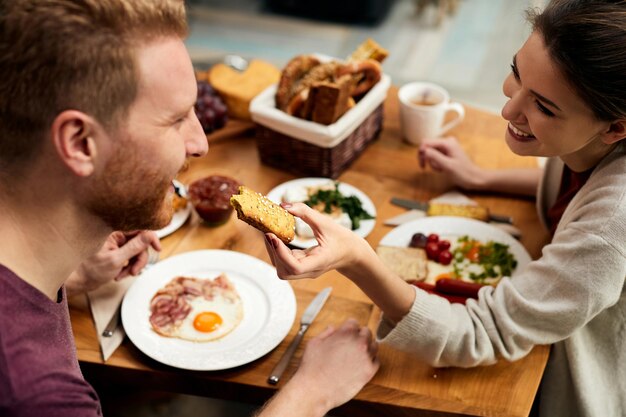 Mujer feliz alimentando a su novio durante el desayuno en casa