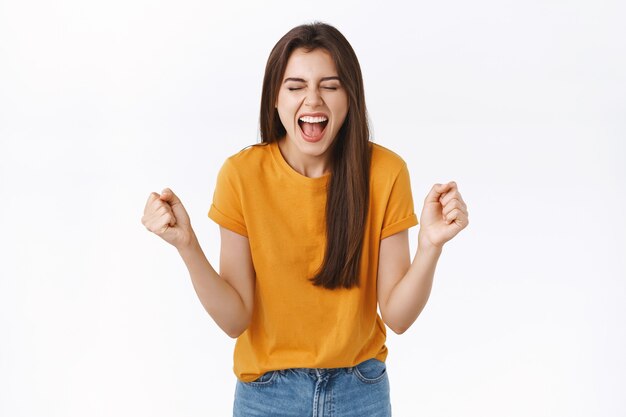 Mujer feliz y aliviada que se regocija con una camiseta amarilla, aprieta los puños y sonríe con alegría, logra el éxito, tiene la suerte de ganar el premio, se convierte en campeona, cierra los ojos y grita satisfecho, triunfando