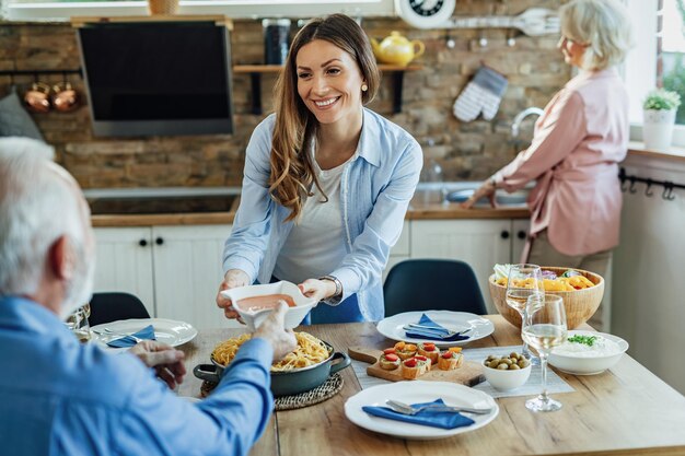 Mujer feliz almorzando con sus padres mayores y trayendo comida a la mesa en el comedor.