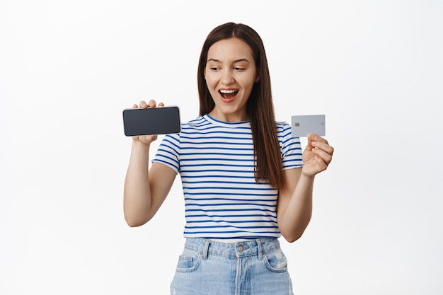 La mujer feliz de los años 20 muestra la pantalla horizontal del teléfono móvil con tarjeta de crédito, riendo y sonriendo, la chica se ve emocionada con el teléfono inteligente, de pie contra el fondo blanco