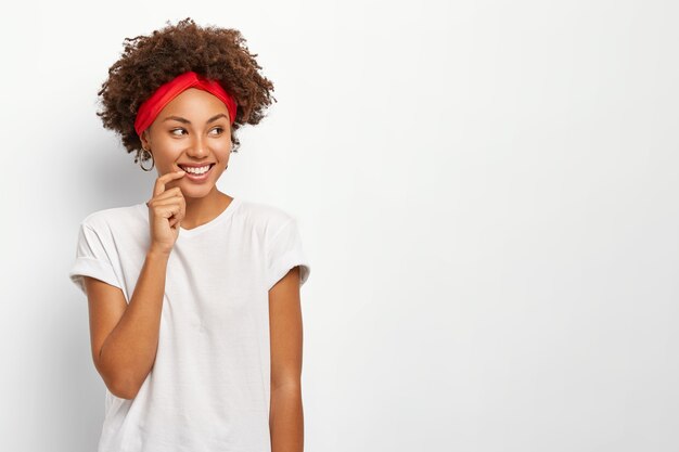 Mujer feliz con apariencia encantadora, mira con alegría a un lado, usa diadema y camiseta casual, tiene dientes perfectos y uniformes