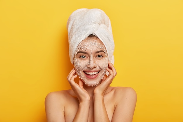 Foto gratuita mujer feliz de aspecto agradable destapa los poros, hace un paso de belleza para mejorar la piel, usa un exfoliante facial nutritivo, mejora la tez, mantiene las manos en las mejillas