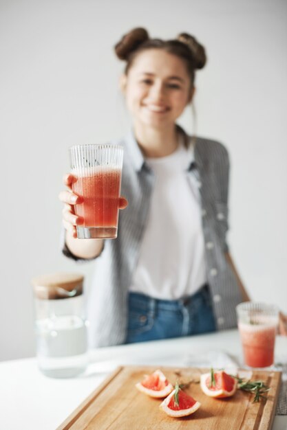 Mujer feliz con los bollos que sonríen estirando el batido de desintoxicación del pomelo sobre la pared blanca. Comida de dieta saludable. Se centran en el vidrio.