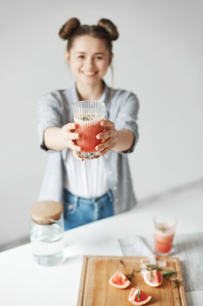 Mujer feliz con los bollos que sonríen estirando el batido de desintoxicación del pomelo sobre la pared blanca. Comida de dieta saludable. Se centran en el vidrio.