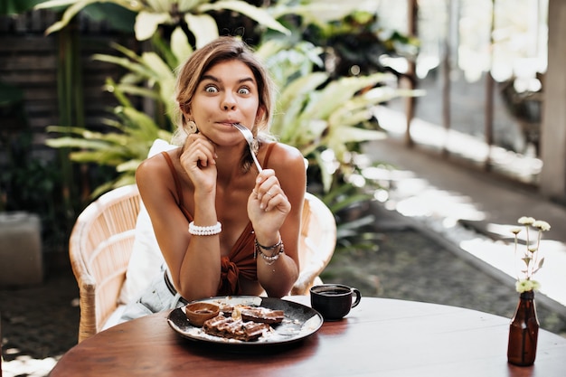 Mujer feliz bronceada en sujetador marrón parece sorprendida, sostiene un tenedor y hace una mueca