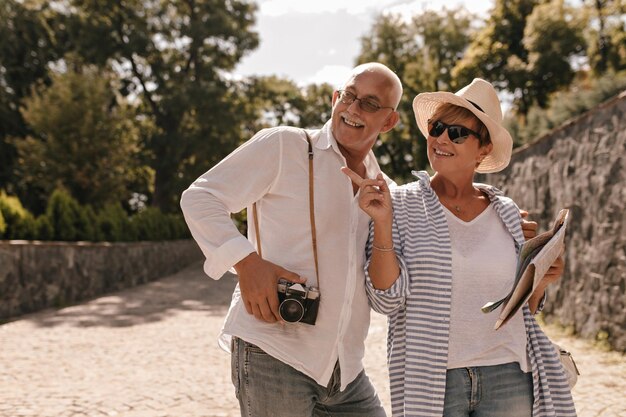 Mujer feliz con cabello corto rubio con gafas de sol y blusa azul con mapa en las manos apunta a un lado y sonriendo con un hombre con camisa blanca con cámara en el parque