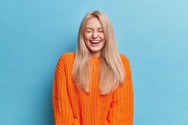 Mujer feliz con cabello largo rubio ríe positivamente escucha algo gracioso cierra los ojos muestra dientes blancos viste suéter de punto naranja