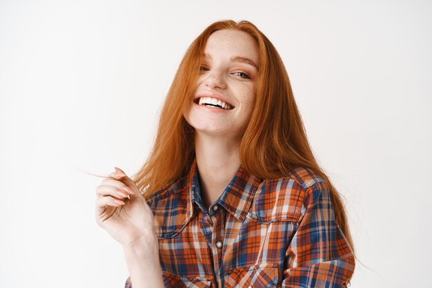 Mujer feliz con cabello pelirrojo, sonriendo y mirando al frente, piel pálida con cara limpia sin maquillaje, pared blanca