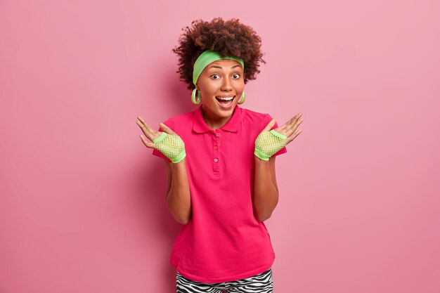 Mujer feliz con cabello rizado natural, levanta las palmas de las manos, se siente confundida, se ríe positivamente, usa guantes deportivos, camiseta rosa brillante, posa en interiores