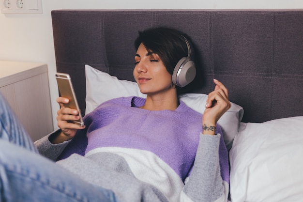 Mujer feliz en casa en una cómoda cama vistiendo suéter de ropa de abrigo, escuchar música